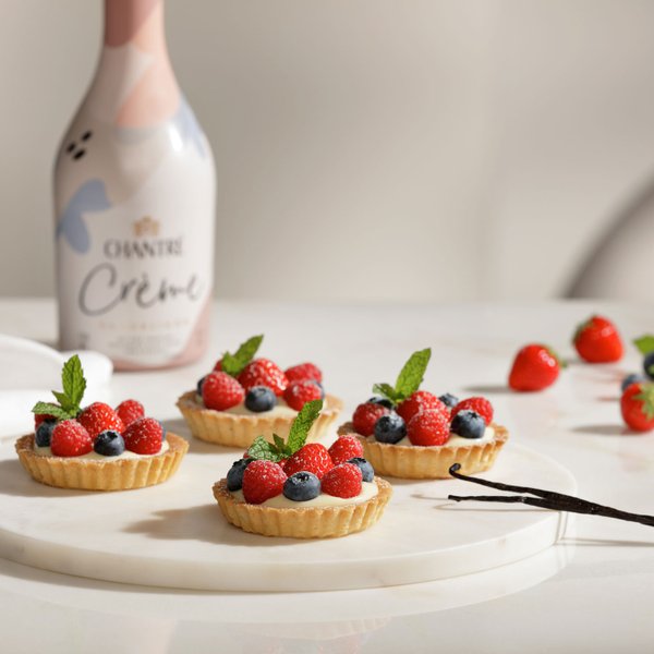 Ein elegantes Arrangement von Obsttörtchen, umgeben von frischen Beeren und einer Flasche Chantre Crème im Hintergrund.