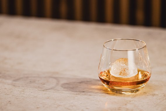 Das Glas mit einem runden Eiswürfel enthält ein bernsteinfarbenes Getränk und steht auf einem minimalistischen Tisch.