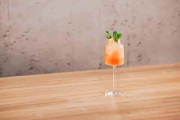 Ein elegantes Glas mit einem erfrischenden, orangefarbenen Cocktail steht auf einem hellen Holztisch.