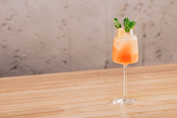 Ein elegantes Glas mit einem erfrischenden, orangefarbenen Cocktail steht auf einem hellen Holztisch.
