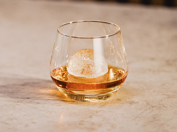 Das Glas mit einem runden Eiswürfel enthält ein bernsteinfarbenes Getränk und steht auf einem minimalistischen Tisch.