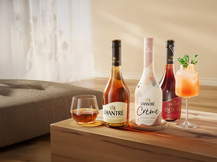 Drei Flaschen Chantré-Likör und ein Glas mit einem fruchtigen Cocktail stehen auf einem Tisch in einem hellen, einladenden Raum.