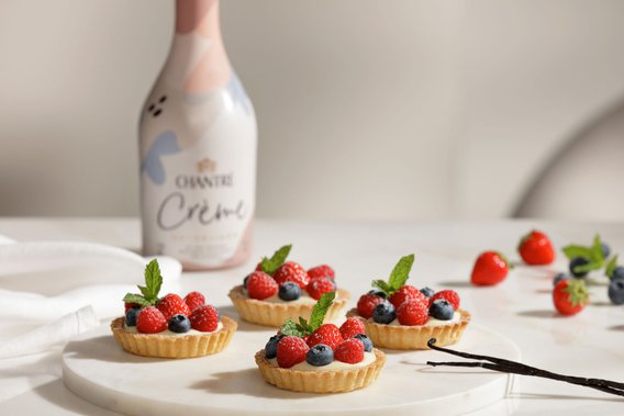 Ein elegantes Arrangement von Obsttörtchen, umgeben von frischen Beeren und einer Flasche Chantre Crème im Hintergrund.