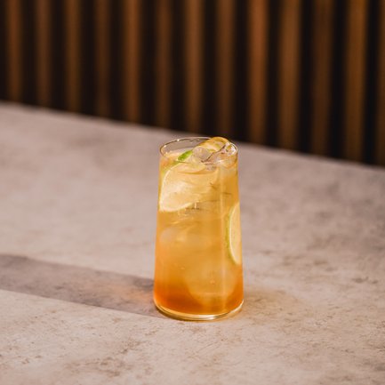 Ein erfrischendes Getränk in einem hohen Glas, gefüllt mit Eiswürfeln und einer Limettenscheibe, steht auf einem hellen Tisch.