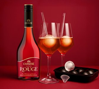 Ein stilvoll arrangiertes Genuss-Set mit einer Flasche Chantré Cuvée Rouge, zwei eleganten Gläsern mit roten Aperitifgetränken, gläsernen Strohhalmen und einer Eiswürfelform mit großen Kugelformen – inszeniert vor rotem Hintergrund.