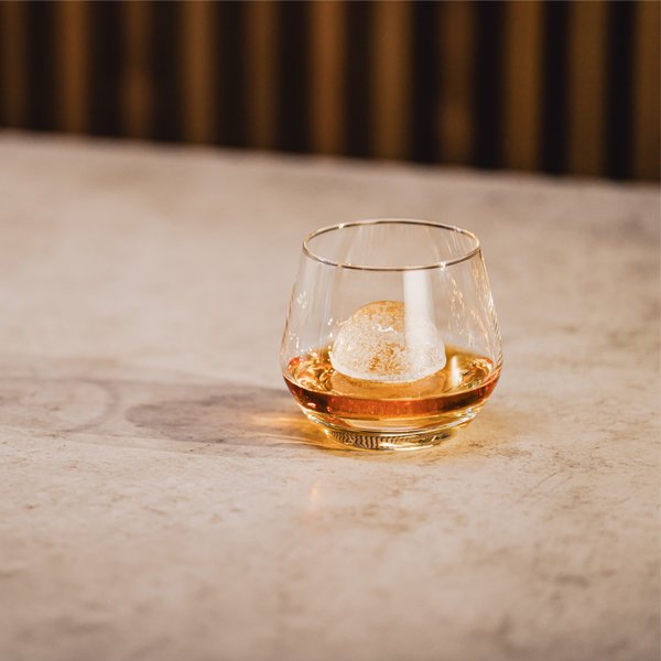 Das Glas mit einem runden Eiswürfel enthält ein bernsteinfarbenes Getränk und steht auf einem minimalistischen Tisch.