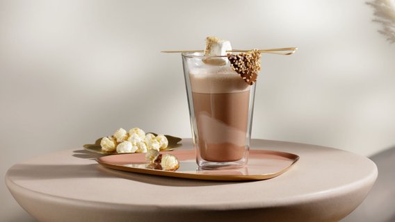 Ein elegantes Glas mit heißer Schokolade, garniert mit einem Marshmallow und Schokolade, steht auf einem runden Tisch.
