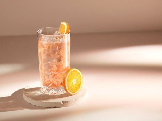 Ein Glas mit einem rosafarbenen Getränk, gefüllt mit Eis, steht auf einem runden, hellen Untersetzer neben einer frischen Orangenscheibe.