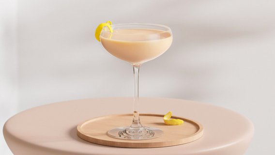 Ein elegantes Cocktailglas mit einer cremigen, hellen Flüssigkeit und einer Zitronenschale auf einem Holztablett.