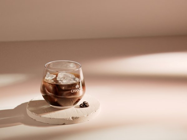 Ein elegantes Glas mit Eiswürfeln und dunklem Getränk steht auf einem runden, hellen Untersetzer, umgeben von sanften Schatten.