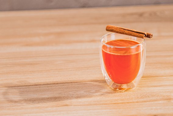 Ein transparentes Glas mit einem warmen, orangefarbenen Getränk, garniert mit einem Zimtstängel auf einem Holzuntergrund.