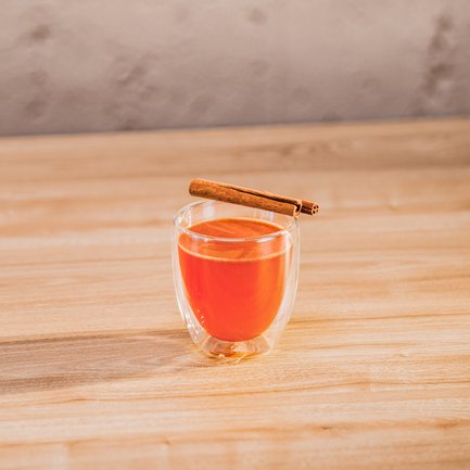 Ein transparentes Glas mit einem warmen, orangefarbenen Getränk, garniert mit einem Zimtstängel auf einem Holzuntergrund.