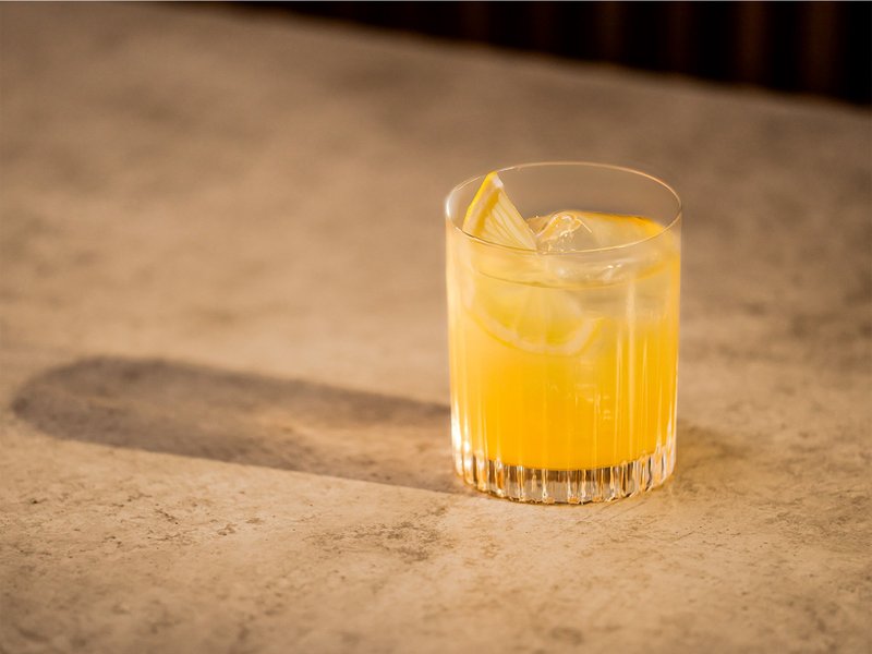 Auf einer glatten, hellen Oberfläche steht ein Cocktailglas mit einem gelben Getränk und einer Zitronenscheibe als Garnitur.