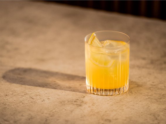 Auf einer glatten, hellen Oberfläche steht ein Cocktailglas mit einem gelben Getränk und einer Zitronenscheibe als Garnitur.
