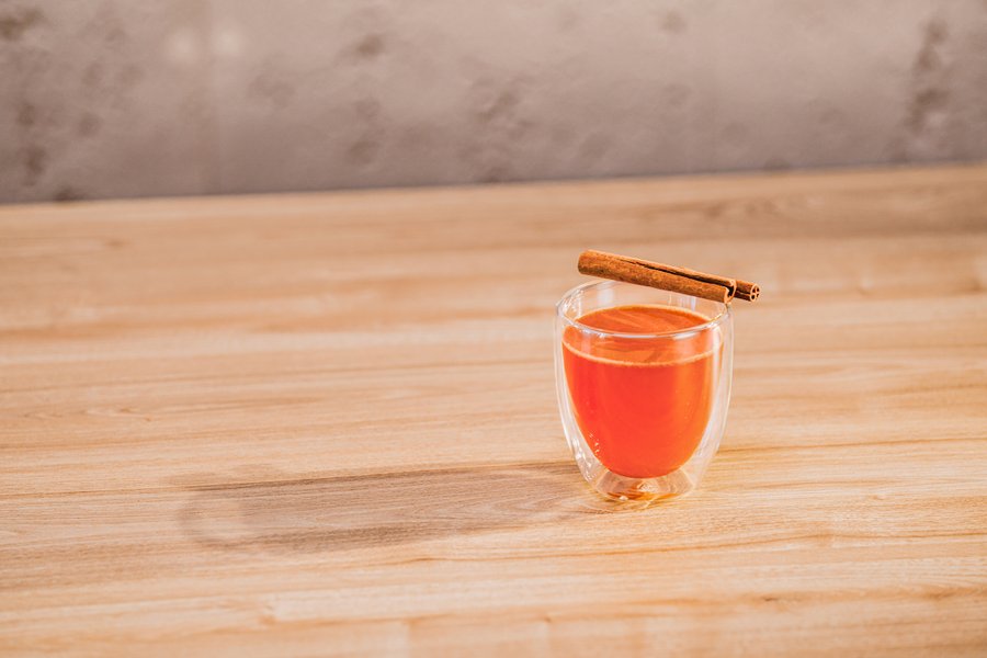 Ein transparentes Glas mit einem warmen, orangefarbenen Getränk, garniert mit einem Zimtstängel auf einem Holzuntergrund.