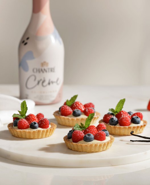 Ein elegantes Arrangement von Obsttörtchen, umgeben von frischen Beeren und einer Flasche Chantre Crème im Hintergrund.