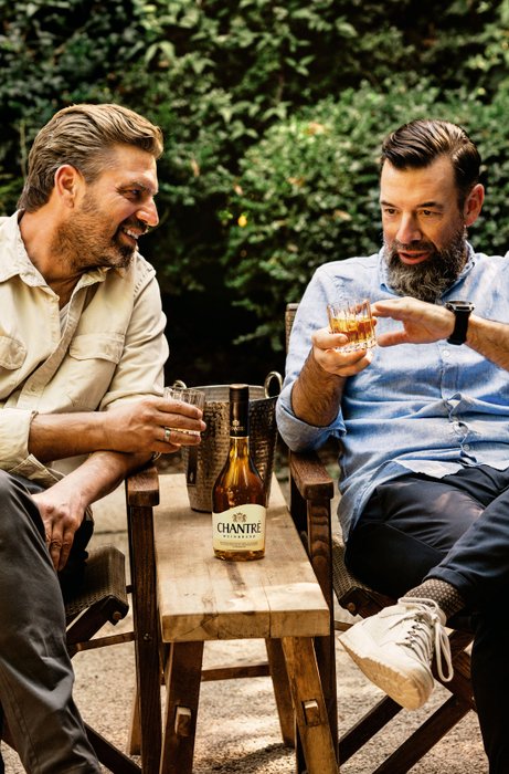 Zwei Männer sitzen entspannt in Gartenstühlen und genießen Drinks, während sie sich angeregt unterhalten.