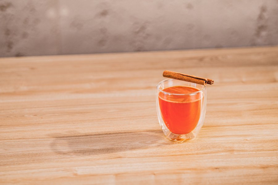 Ein transparentes Glas mit einem warmen, orangefarbenen Getränk, garniert mit einem Zimtstängel auf einem Holzuntergrund.