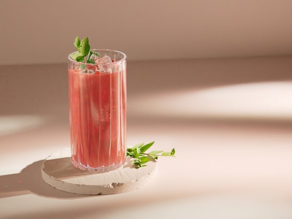 Ein stilvolles Glas mit einem pinken Cocktail, das auf einem runden, hellen Untersetzer steht, umgeben von sanften Lichtschatten.