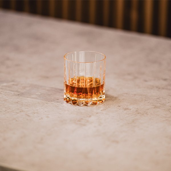 Ein elegantes Glas mit bernsteinfarbenem Getränk steht auf einer hellen, strukturierten Oberfläche.