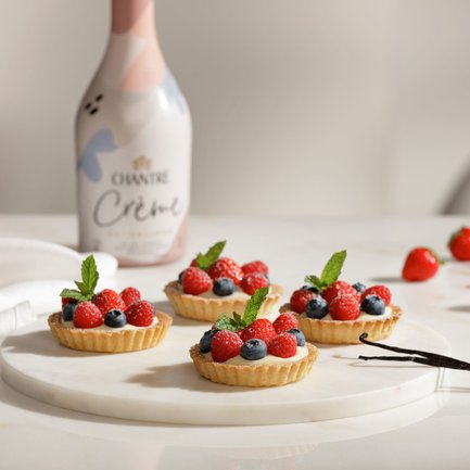 Ein elegantes Arrangement von Obsttörtchen, umgeben von frischen Beeren und einer Flasche Chantre Crème im Hintergrund.