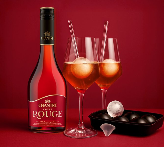 Ein stilvoll arrangiertes Genuss-Set mit einer Flasche Chantré Cuvée Rouge, zwei eleganten Gläsern mit roten Aperitifgetränken, gläsernen Strohhalmen und einer Eiswürfelform mit großen Kugelformen – inszeniert vor rotem Hintergrund.