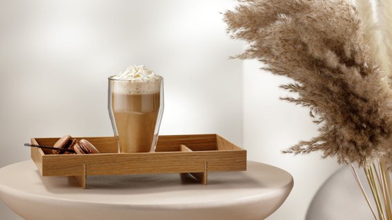 Ein Glas mit einem cremigen Getränk, garniert mit Sahne, steht auf einem Holztablett neben köstlichen Macarons.