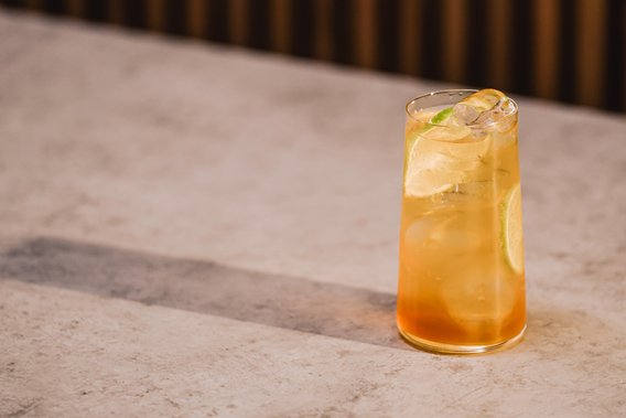 Ein erfrischendes Getränk in einem hohen Glas, gefüllt mit Eiswürfeln und einer Limettenscheibe, steht auf einem hellen Tisch.