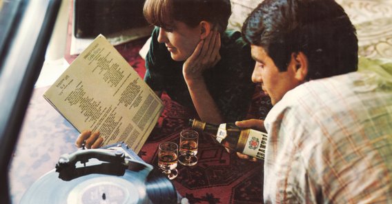 Eine Frau und ein Mann sitzen auf einem Teppich, während sie eine Schallplatte anschauen und ein Chantré Getränk genießen.