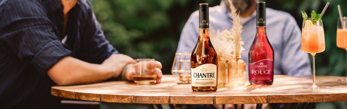 Auf einem Holztisch stehen zwei Flaschen CHANTRÉ Weinbrand und Cocktails, während zwei Männer entspannt miteinander plaudern.
