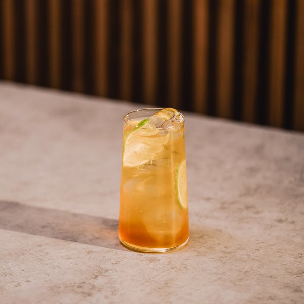 Ein erfrischendes Getränk in einem hohen Glas, gefüllt mit Eiswürfeln und einer Limettenscheibe, steht auf einem hellen Tisch.