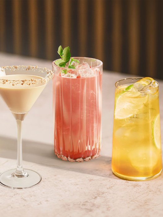 Drei verschiedene Cocktails stehen auf einem hellen Tisch, jeder in einem einzigartigen Glas mit Eis und frischen Zutaten.