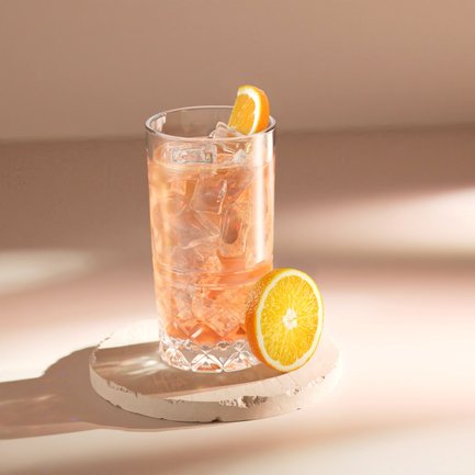 Ein Glas mit einem rosafarbenen Getränk, gefüllt mit Eis, steht auf einem runden, hellen Untersetzer neben einer frischen Orangenscheibe.